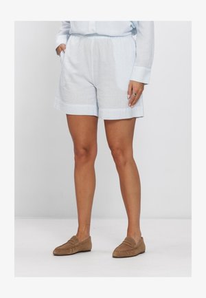 Personne portant un short blanc texturé avec des poches, un haut à manches longues assorti et des mocassins marron à enfiler, debout devant un fond uni.