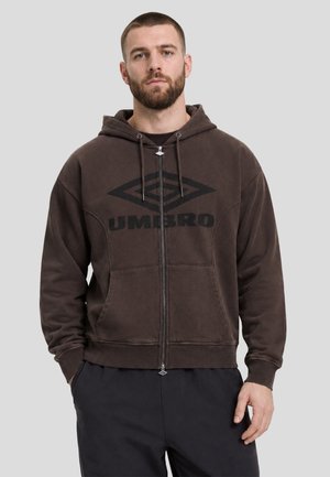Hombre con cabello corto y barba que lleva una sudadera marrón con capucha y cremallera, con logo negro de Umbro, de pie frente a un fondo gris liso.