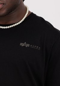 Černé bavlněné tričko s jemným šedým logem "ALPHA INDUSTRIES". Hladký povrch a klasický design s kulatým výstřihem. Akcent v podobě perlového náhrdelníku.