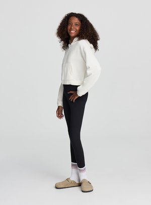 Giovane donna con capelli ricci che indossa una felpa corta bianca con cappuccio, leggings neri, calzini bianchi a righe e scarpe slip-on beige, in piedi di lato.