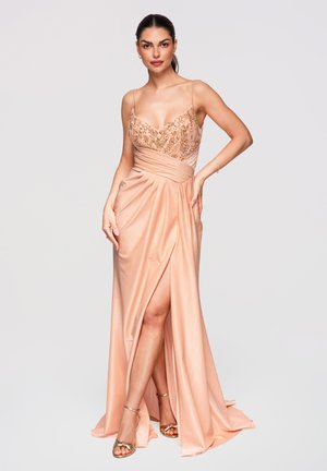 Femme en robe satinée pêche avec corsage orné, fente haute jusqu'à la cuisse et talons dorés, debout devant un fond uni.