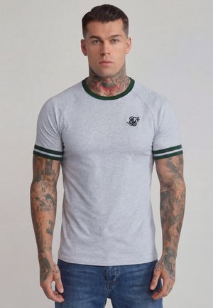 Man with tattoos on neck and arms wearing a light gray fitted T-shirt with green trim and logo on chest, paired with blue jeans.