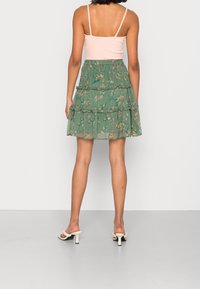 Falda verde de volantes con patrón floral, hecha de tela ligera. Combinada con una blusa rosa claro y sandalias blancas de tacón.