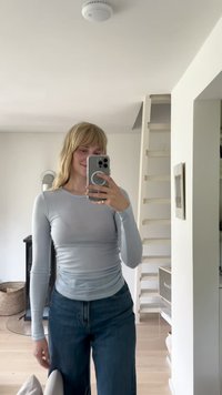 Vaaleansininen pitkähihaine paita, jossa on vartalonmyötäinen muotoilu ja pyöreä pääntie, yhdistettynä rentoihin tummansinisiin farkkuihin. Moderni sisustus taustalla.