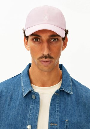 Uomo con baffi che indossa un berretto rosa chiaro, una giacca di jeans blu e una camicia bianca, guarda dritto davanti a uno sfondo bianco.
