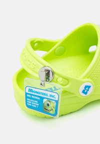 Dzelteni-zaļi Crocs ar fakturētu virsmu, ar siksniņu un plastmasas insigniju, uz kuras attēlots varonis no "Monstru korporācijas".