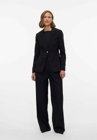 Completo giacca nera e pantaloni, vestibilità su misura con chiusura a un bottone e tessuto testurizzato. Il modello è in piedi contro uno sfondo bianco semplice.