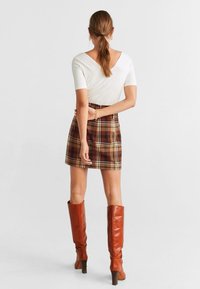 Top blanco de manga corta, falda corta de cuadros marrones y botas de cuero marrón hasta la rodilla con un tacón grueso, vistas desde atrás.