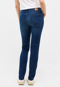 Jeans din denim de un albastru închis, cu un croi drept, buzunare spate și o etichetă din piele maro la talie. Textură netedă, detalii minime.