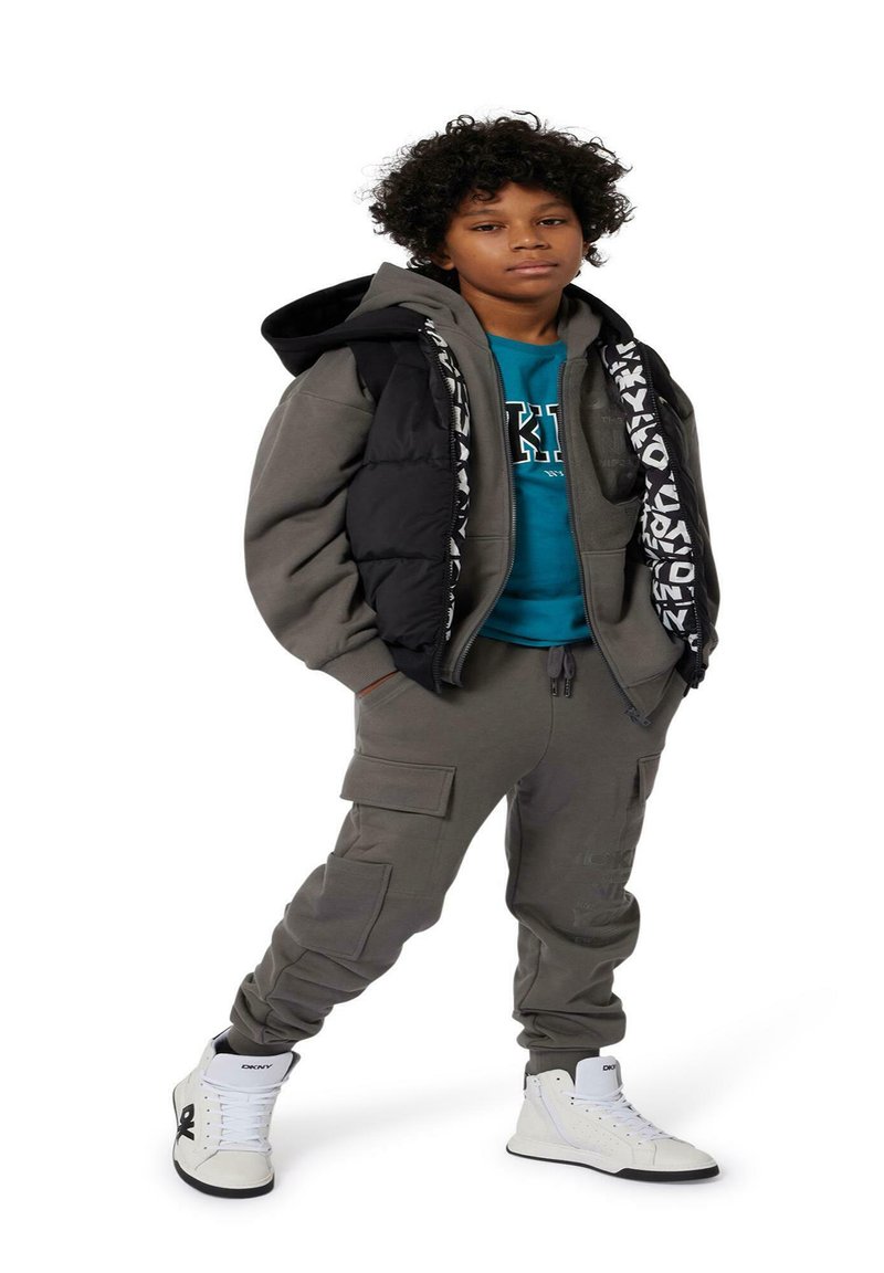 Survêtement gris avec un gilet matelassé noir, présentant un accent à rayures à motifs. Chemise bleue en dessous et baskets montantes blanches.