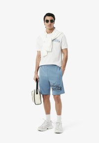Camisa polo blanca ligera con logo azul, combinada con pantalones cortos azules con un estampado negro. Zapatillas blancas y un bolso crema con acentos negros.