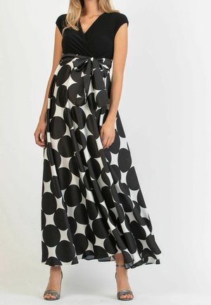Kobieta ubrana w czarny top i czarno-białą spódnicę maxi w groszki zawiązaną w talii, stojąca z rozluźnionymi ramionami.