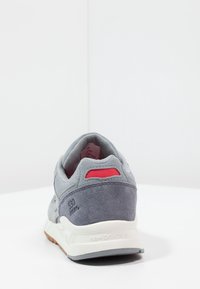 Grijze New Balance 530 sneaker gezien vanaf de achterkant, met een uitzicht op de suède textuur, witte zool en rode stoffen details bij de hielkraag.