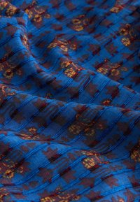 Tissu bleu avec des motifs floraux et géométriques bordeaux et jaunes, présenté en plis doux mettant en valeur la texture et les couleurs vives.