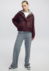 Cazadora burdeos con una parte superior texturizada y una sección inferior lisa, que presenta una capucha gris y un bolsillo. Pantalones de pierna ancha grises con una textura sutil.