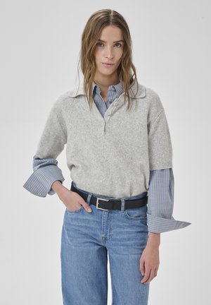 Młoda kobieta ubrana w szary sweter na warstwach, nałożony na niebieską koszulę w paski z podwiniętymi mankietami, niebieskie jeansy i czarny pasek, stoi na tle o neutralnym kolorze.