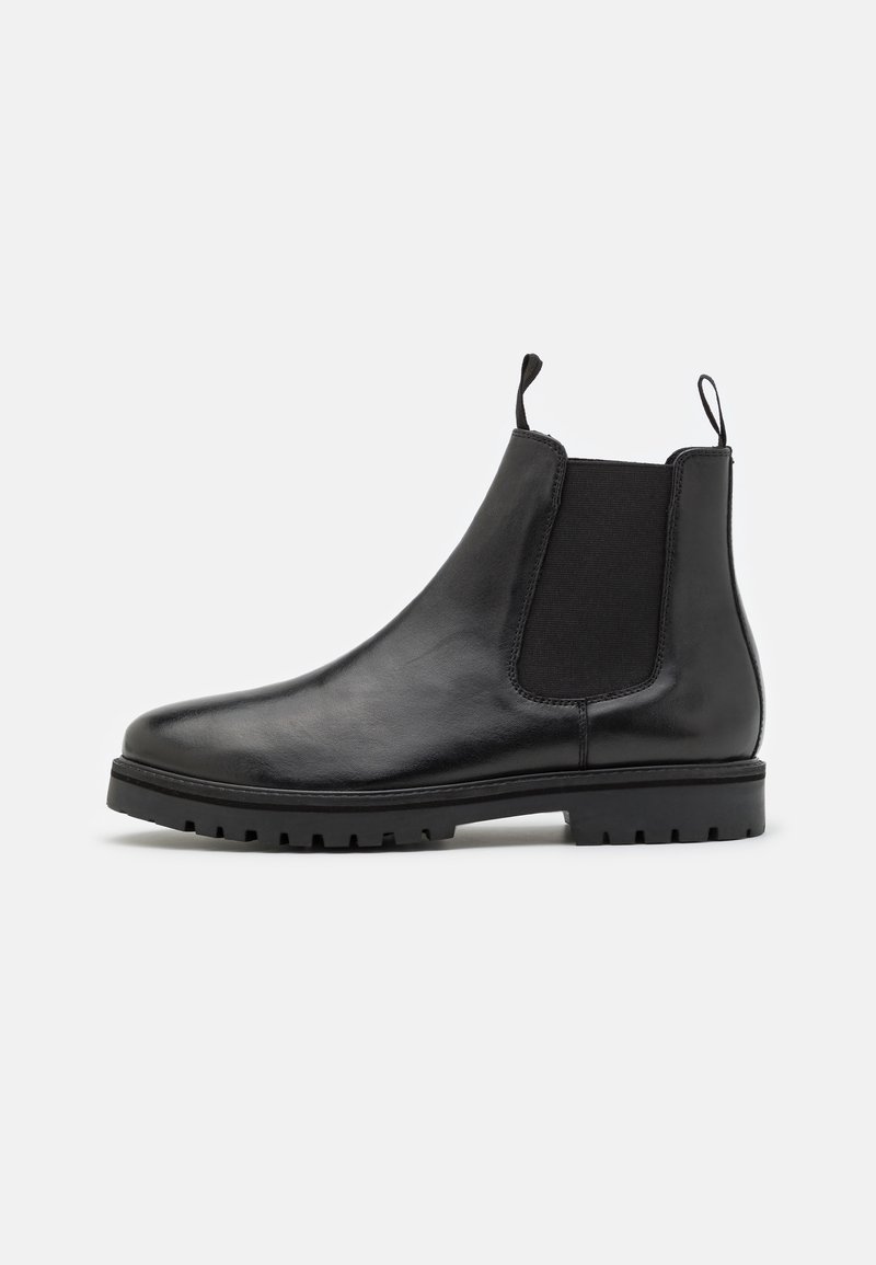 Bota Chelsea de cuero negro con paneles elásticos en los laterales, pestañas de agarre en la parte delantera y trasera, y suela de goma gruesa sobre fondo blanco.