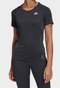 Žena oblečená v těsně padnoucím černém krátkém funkčním tričku Adidas a odpovídajících černých legínách, stojí s uvolněnýma rukama podél těla.