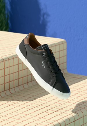 Sort sneaker med brun krave og hvid sål, hvilende på cremefarvede trin med røde gitterlinjer, mod en blå væg som baggrund.