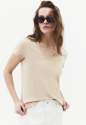 Vrouw met lang haar die zwarte zonnebril draagt, een beige kortärmelig V-hals top en witte broek en haar shirtzoom verstelt.