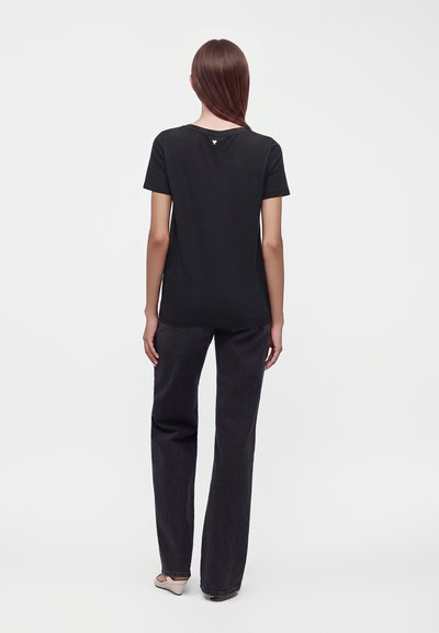 Femme aux longs cheveux bruns de dos, portant un t-shirt noir à manches courtes, un jean sombre à jambes larges et des chaussures plates de couleur claire, debout sur fond blanc.