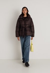 Jakna s punim kaputom u tamnosmeđoj boji sa pojasom u struku, visokim ovratnikom i kontrastnom žutom torbom. Nosena uz svijetloplave hlače širom nogavica i crne cipele.