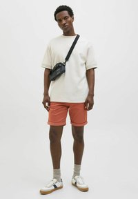 Men's beige short-sleeve T-shirt, coral shorts with rolled cuffs, white sneakers with navy accents, and a small black crossbody bag.