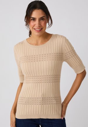 Frau lächelt, trägt ein beiges kurzärmliges Strickoberteil mit gerippten und gemusterten horizontalen Reihen, kombiniert mit dunkelblauen Jeans.