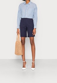 Camicia azzurra a righe con bottoni, pantaloni corti blu navy, scarpe con tacco alte bianche e una borsa a tracolla intrecciata beige. Design semplice e strutturato.