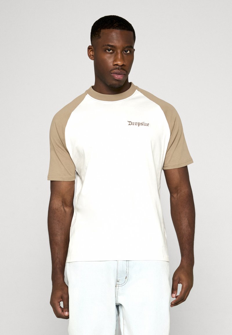 Homme portant un t-shirt raglan à manches courtes blanc et beige avec le texte "Dropsike" sur la poitrine, associé à un pantalon clair, debout.