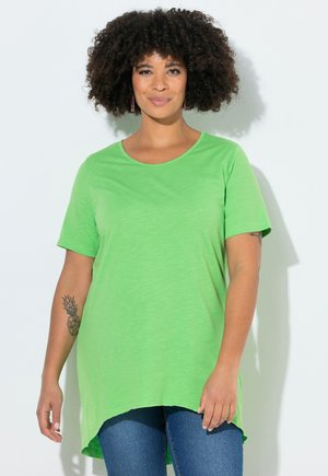 Woman with curly hair wearing a bright green short-sleeve shirt and blue jeans, standing against a white background.