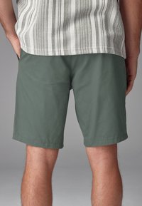 Pantaloni verdi realizzati in tessuto leggero, caratterizzati da un design semplice con taglio dritto e orlo cucito, abbinati a una camicia a righe.