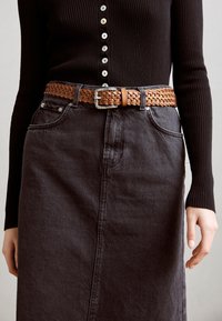 Vävd brun läderbälte med en silverspänne som bärs över en svart ribbad långärmad tröja och en mörkgrå denimkjol, vilket framhäver texturer och färger.