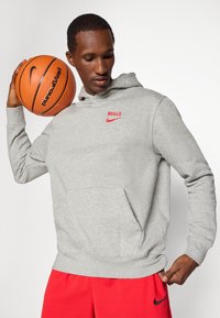 Grå hættetrøje med en frontlomme, der har røde "BULLS" og Nike-logoer. Bæreren holder en orange basketball, iført røde shorts.