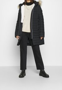 Svart puffig jacka med huva kantad med päls. Har en quiltad design, dragkedja och vit, mjuk foder. Matchas med en krämfärgad tröja och svarta byxor.