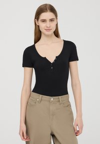 Body corto nero a maniche corte con scollatura a V profonda e patta con tre bottoni; tessuto morbido, vestibilità aderente, abbinato a pantaloni beige chiaro.