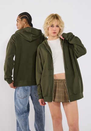 Olive green hooded sweatshirt with a zip front, relaxed fit, and ribbed cuffs. Model wears it over a cropped white top and plaid shorts.