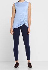 Ljusblå ärmlös topp med en vriden detalj vid midjan, i kombination med marinblå leggings och svarta träningsskor. Slät tygkvalitet och åtsittande design.