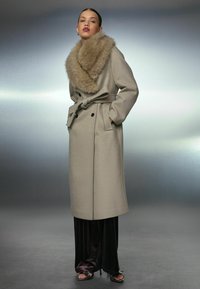 Beige wollen overjas met een grote faux fur kraag, dubbelgeknoopt front, een strik taille en lange mouwen. Gecombineerd met donkere satijnen broek en sandalen.