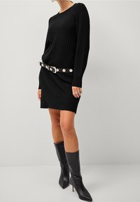 Robe-pull noire côtelée à manches longues, accessoirisée d'une large ceinture noire avec des accents circulaires métalliques et des bottes noires montantes jusqu'aux genoux.