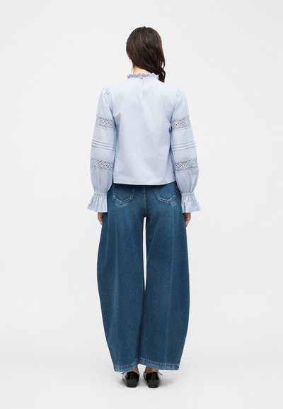 Γυναίκα που φορά ανοιχτό μπλε μπλούζα με δαντελένιες λεπτομέρειες και μπλε τζιν παντελόνι με φαρδιά μπατζάκια, στέκεται γυρισμένη προς τα πίσω σε λευκό απλό φόντο.