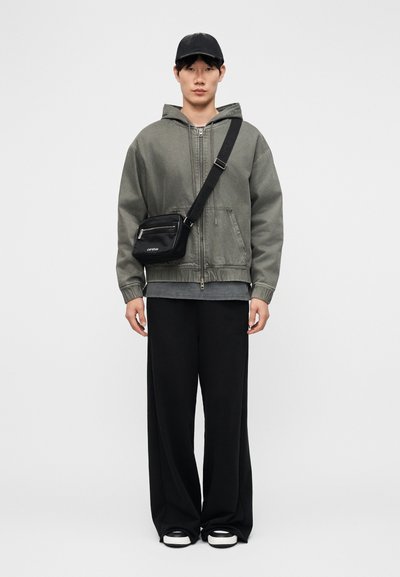 Νέος άνδρας με ξεθωριασμένο πράσινο φουτερζάκι με φερμουάρ, μαύρο παντελόνι φαρδιάς γραμμής, μαύρο καπέλο, μαύρη τσάντα ώμου και ασπρόμαυρα αθλητικά παπούτσια, στέκεται όρθιος.