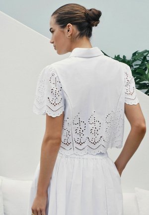 Sieviete ar brūniem matiem sasietiem bulciņā, tērpusies baltā īsroku krekliņā ar izšūtu mežģīņu rakstu un saskanīgā svārku komplektā, stāv uz gaiša fona.