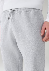 Inimene, kes kannab helehalli spordipükse reguleeritava paelaga vöökohal ja valget pika varrukaga särki, lähedalt vaadatuna.