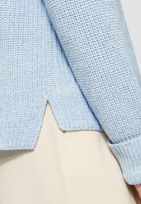 Hellblauer Strickpullover mit strukturiertem Muster, featuring gerippte Bündchen und einen gespaltenen Saum, über einem cremefarbenen Kleidungsstück getragen.