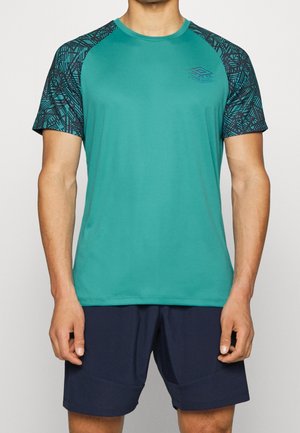 Mand iført en teal kortærmet sportstrøje med mønstrede sorte ærmer, kombineret med mørkeblå atletikshorts, stående mod en ensfarvet baggrund.