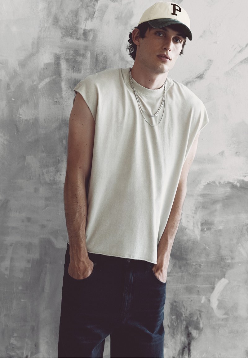 Jeune homme portant un t-shirt blanc sans manches, un pantalon noir, une casquette blanche avec la lettre P, et des chaînes argentées, debout devant un mur gris texturé.