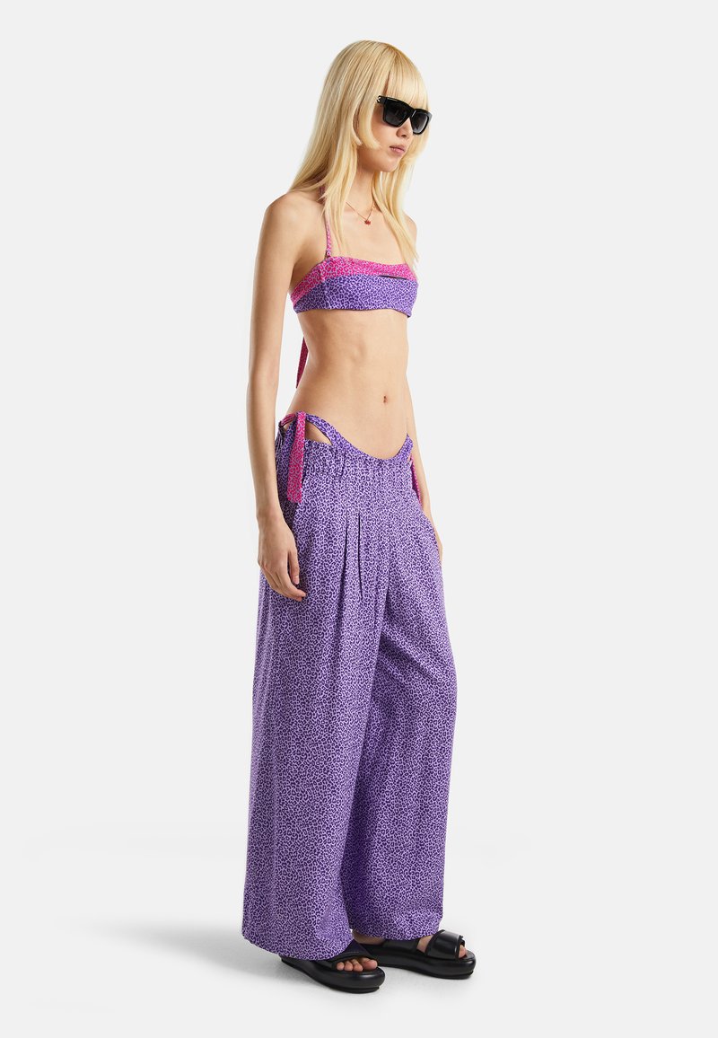 Haut de bikini à motifs violets avec bordures roses et pantalon large violet avec liens sur les côtés, porté avec des lunettes de soleil noires et des sandales à plateforme.