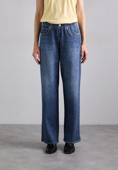Pantalons en denim à jambes larges de couleur bleu moyen, présentant un design classique à cinq poches, un léger délavage et une taille élastique. Associés à des chaussures noires.