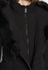 Czarna kurtka puchowa z wysokim kołnierzem, pikowanym materiałem, zapięciem na zamek z przodu oraz sztucznym futrem na ramionach. Teksturowane boczne panele.